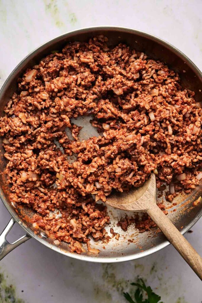 Cooked ground meat mixed with tomato sauce and chopped onions in a skillet, perfect for stuffing into zucchini boats and stirred with a wooden spoon.