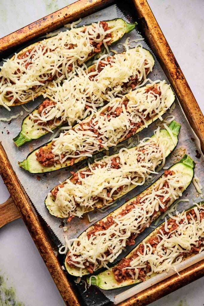 A baking tray with six zucchini boats stuffed with ground meat sauce and topped with shredded cheese, ready to be baked.