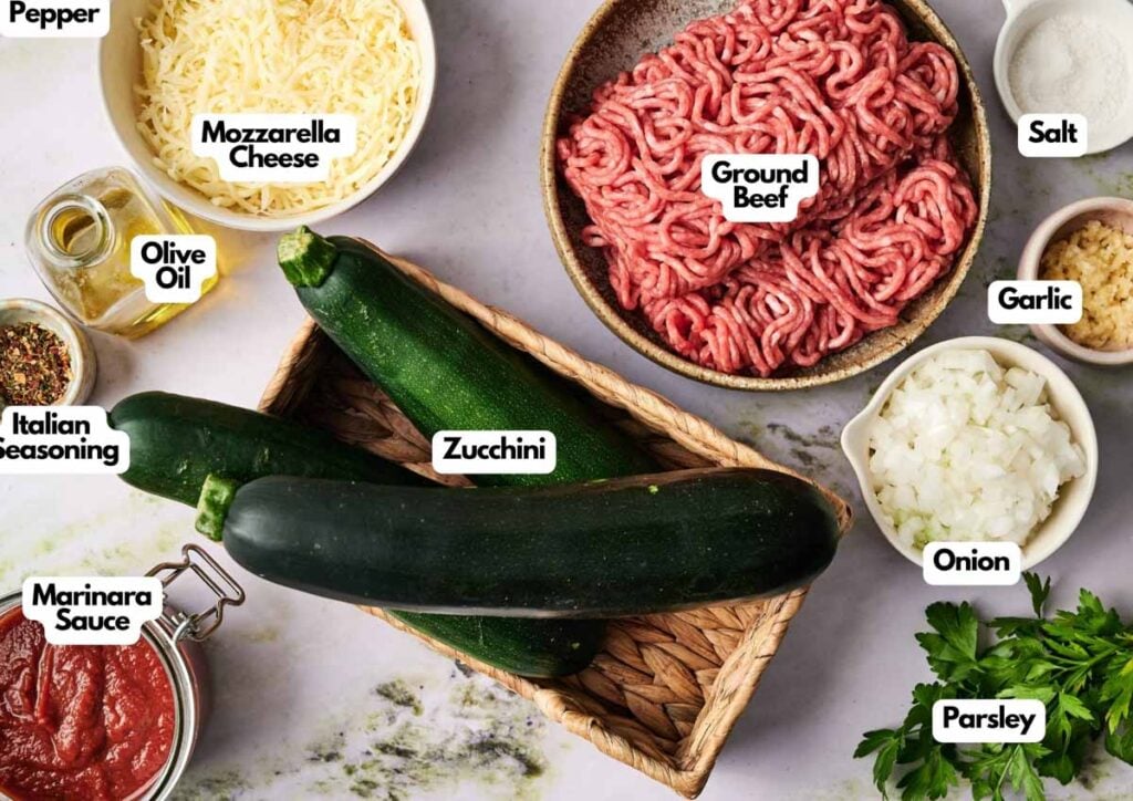 Various labeled ingredients for a zucchini casserole, similar to those used in zucchini boats, are arranged on a table, including ground beef, zucchini, mozzarella cheese, marinara sauce, and seasonings.