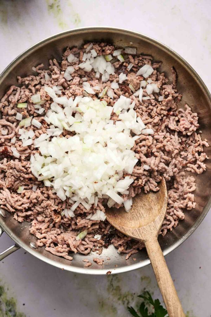 Chopped onions are added to browned ground beef in a skillet, with a wooden spoon resting inside the pan—perfect for stuffing zucchini boats.