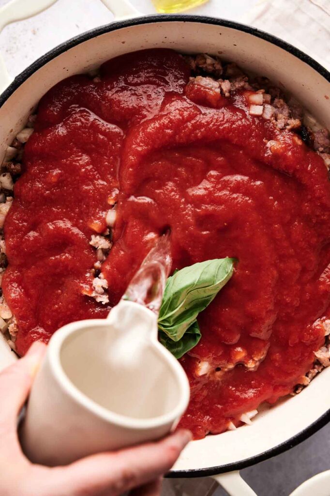A hand pours water from a cup into a pot containing ground meat, chopped onions, tomato sauce, and a fresh basil sprig&mdash;the perfect start for hearty baked ziti.
