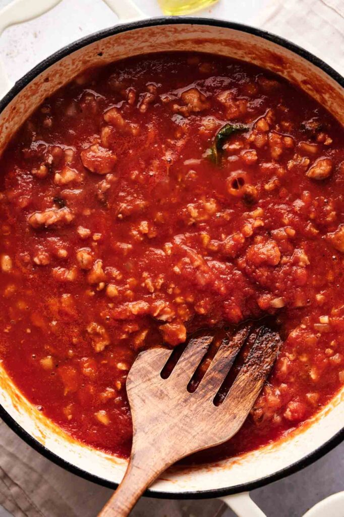 A pot of tomato-based meat sauce with ground meat, perfect for layering into baked ziti, being stirred with a wooden spoon on a white surface.