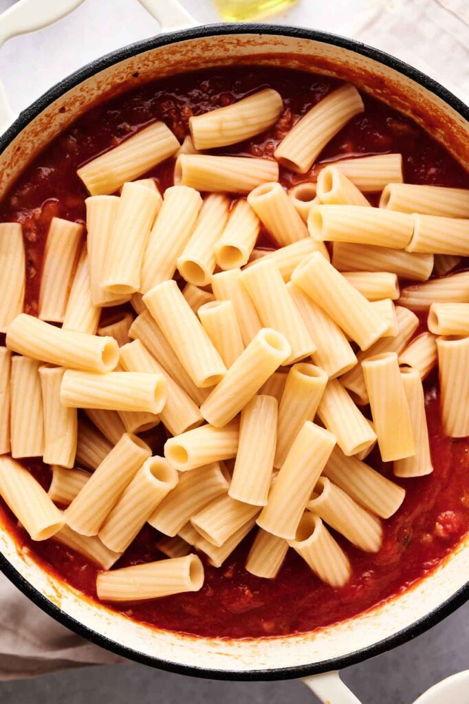 A pot of cooked rigatoni pasta sits on top of rich red tomato sauce, ready to be mixed together for a delicious baked ziti-inspired dish.