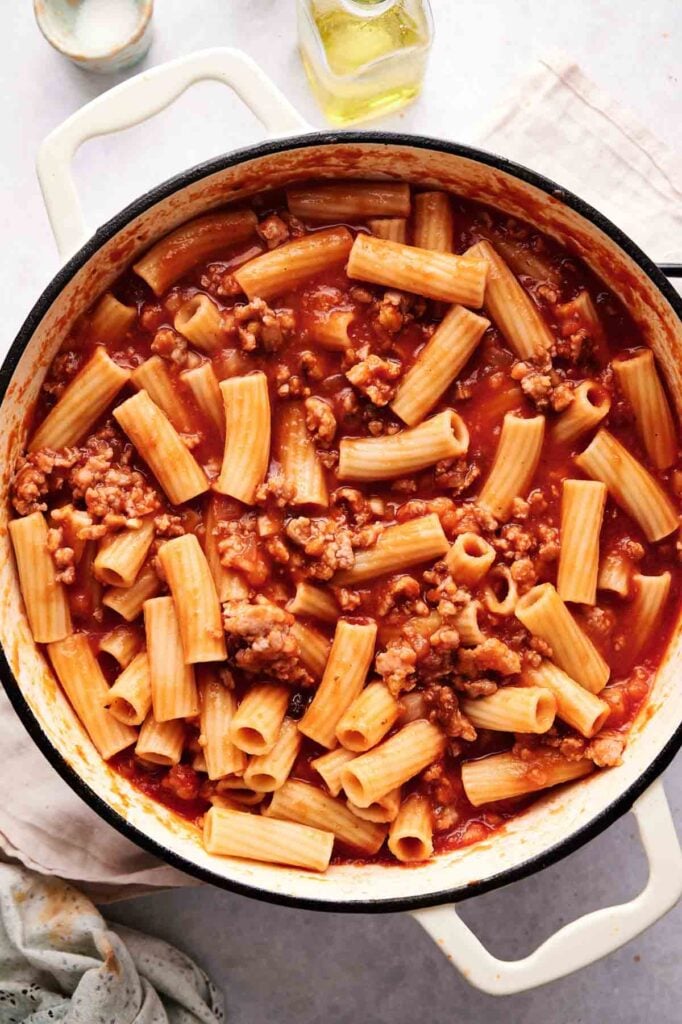 Large pot filled with rigatoni pasta mixed with ground meat and tomato sauce, reminiscent of a classic baked ziti, viewed from above on a light surface.