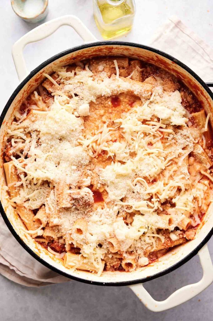 A casserole dish filled with baked ziti, topped with melted cheese and grated parmesan, set on a light-colored surface.