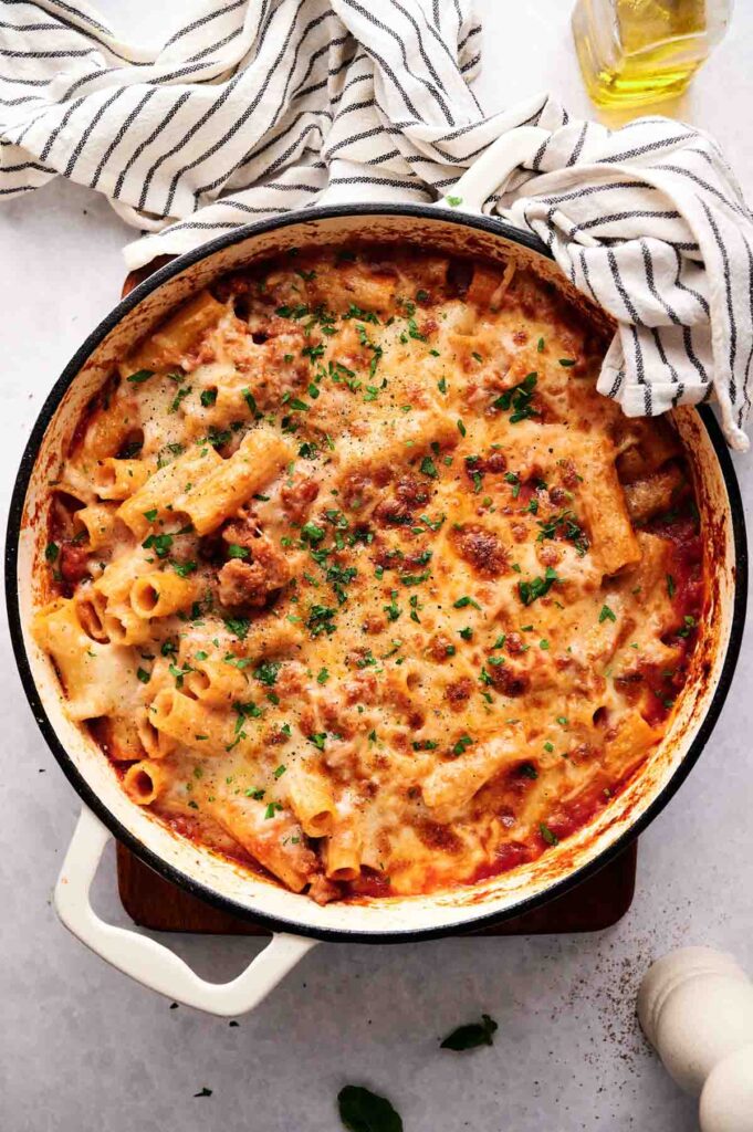 A white casserole dish filled with baked ziti, topped with melted cheese and parsley, sits on a white surface next to a striped towel and a bottle of olive oil.