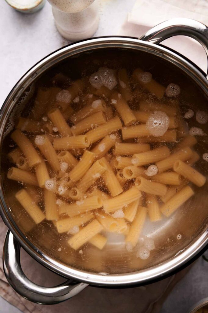 A pot of rigatoni pasta boils in water on the stovetop, with foam forming on top&mdash;perfectly cooked for a classic baked ziti dish.