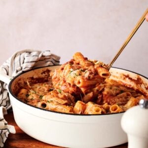 A hand lifts a serving of baked ziti with tomato sauce and melted cheese from a white casserole dish sitting on a wooden board.