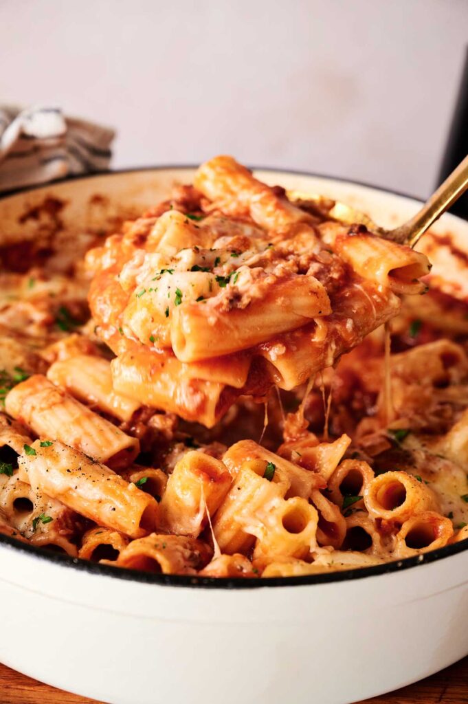 A close-up of baked rigatoni pasta with melted cheese and tomato sauce, reminiscent of classic baked ziti, being served from a white casserole dish.