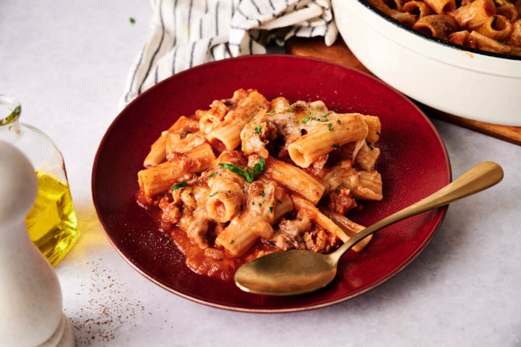 A red plate of baked rigatoni pasta with tomato sauce, ground meat, and melted cheese&mdash;similar to classic baked ziti&mdash;is garnished with herbs and sits next to a gold spoon on a white table.
