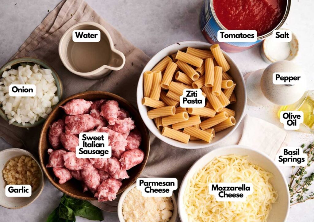 Various labeled ingredients for a baked ziti dish are arranged on a table, including ziti, sweet Italian sausage, onion, garlic, Parmesan, mozzarella, tomatoes, olive oil, basil, salt, pepper, and water.