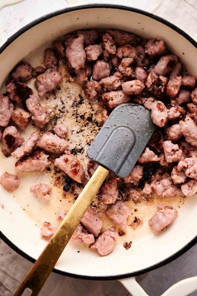 Cooked sausage pieces being stirred in a white pot with a gray spatula, perfect for adding to baked ziti recipes.