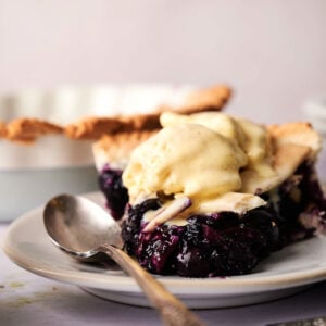 A slice of blueberry pie topped with vanilla ice cream sits on a white plate, a spoon beside it, ready to enjoy.