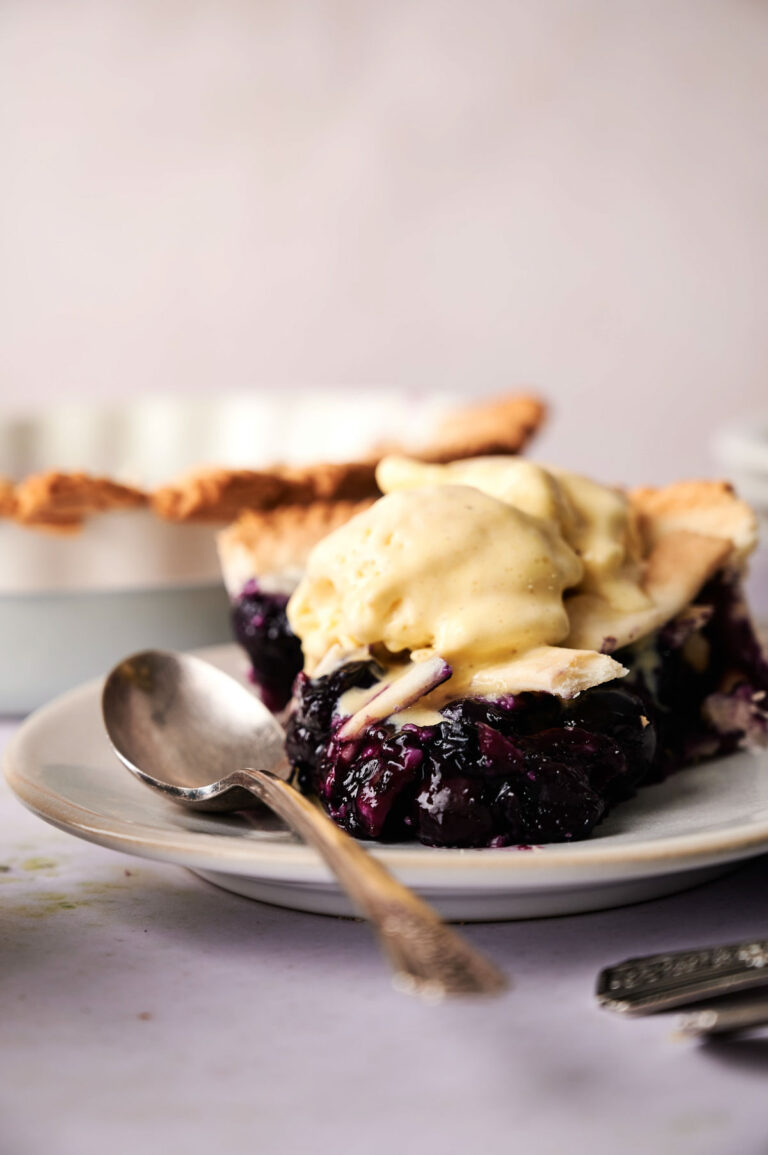 A slice of blueberry pie topped with vanilla ice cream sits on a white plate, a spoon beside it, ready to enjoy.
