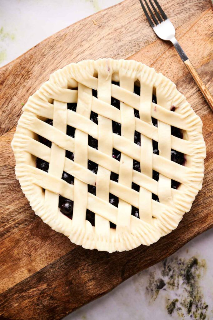 Unbaked blueberry pie with a lattice crust sits on a wooden cutting board next to a fork.