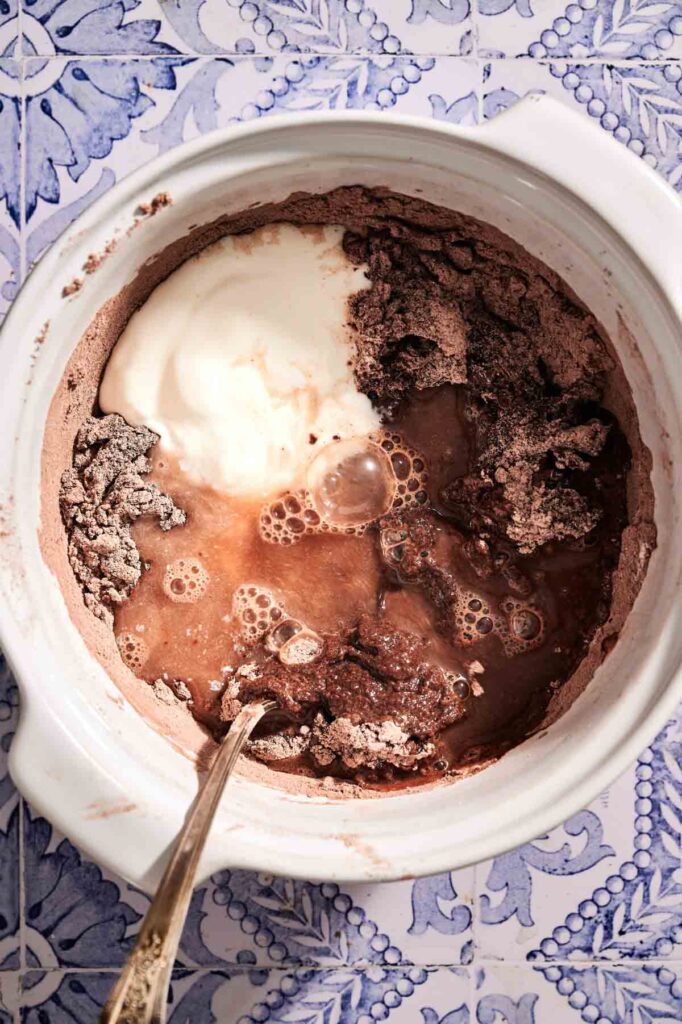 A mixing bowl with cocoa powder, flour, yogurt, and water being combined with a spoon for chocolate chip muffins, placed on a blue and white patterned surface.