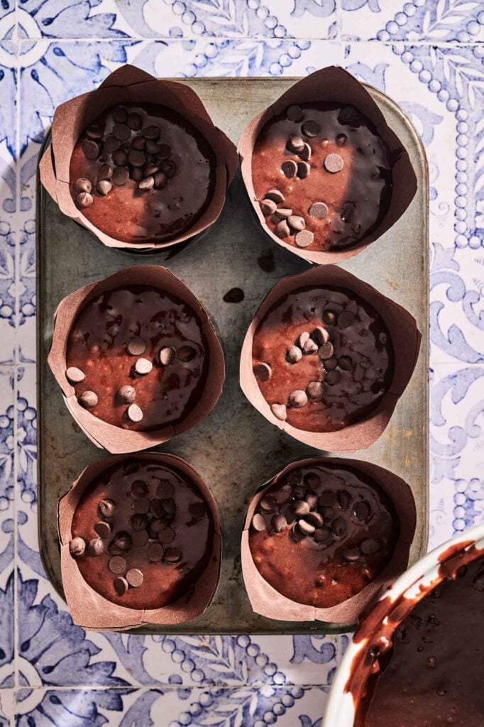 Six unbaked chocolate chip muffins with extra chocolate chips in brown paper liners sit in a baking tray on a patterned surface.