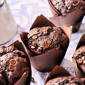 Four delicious chocolate chip muffins topped with extra chocolate chips sit in brown paper wrappers on a patterned surface.