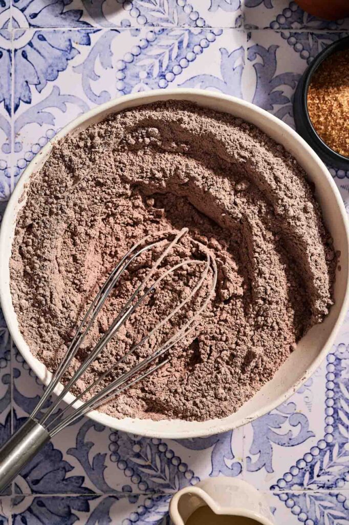 A metal whisk rests in a bowl filled with mixed dry cocoa powder ingredients for chocolate chip muffins on a patterned blue and white surface.