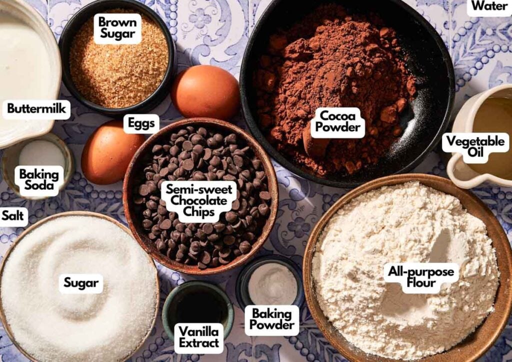Overhead view of labeled baking ingredients for chocolate chip muffins, including flour, cocoa powder, sugar, brown sugar, eggs, buttermilk, oil, water, salt, baking soda, baking powder, chocolate chips, and vanilla.