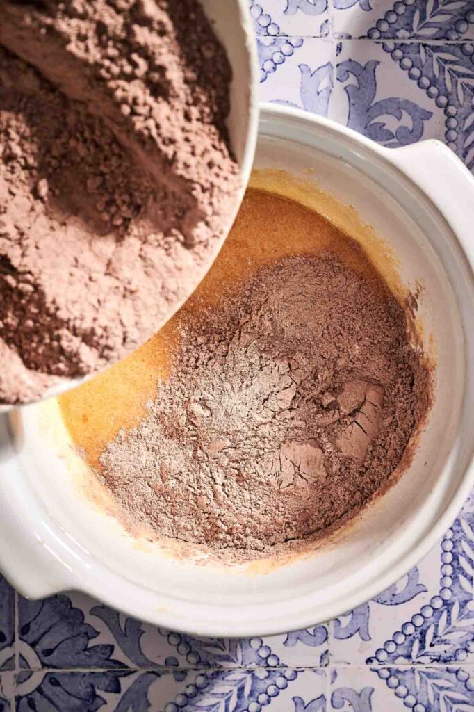 Cocoa powder and flour mixture being poured into a bowl of wet ingredients to make delicious chocolate chip muffins on a blue and white patterned surface.