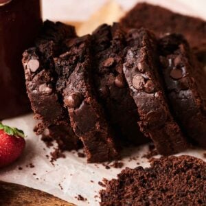 Sliced Chocolate Zucchini Bread loaf with chocolate chips on parchment paper, alongside a strawberry and a mug on a wooden surface.