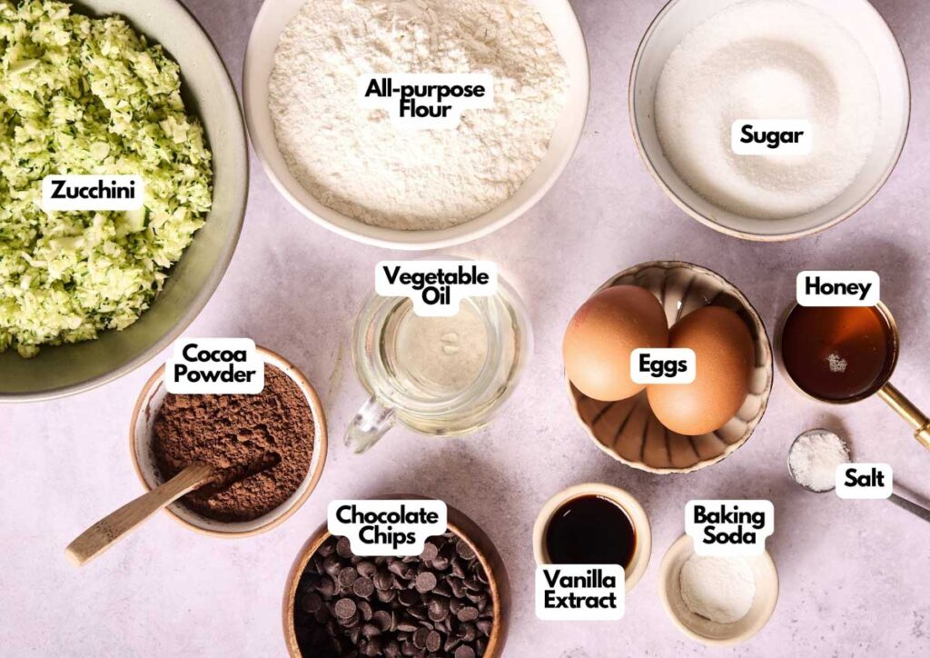 Top-down view of labeled baking ingredients for Chocolate Zucchini Bread in bowls, including zucchini, flour, sugar, cocoa powder, chocolate chips, eggs, honey, vegetable oil, vanilla extract, baking soda, and salt.