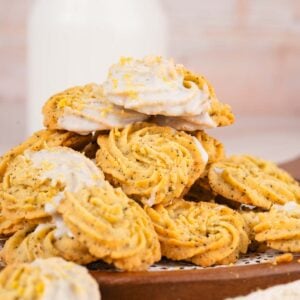 A pile of swirled Earl Grey Lemon Cookies with poppy seeds, white icing, and lemon zest sits on a wooden tray, with a bottle of milk in the background.