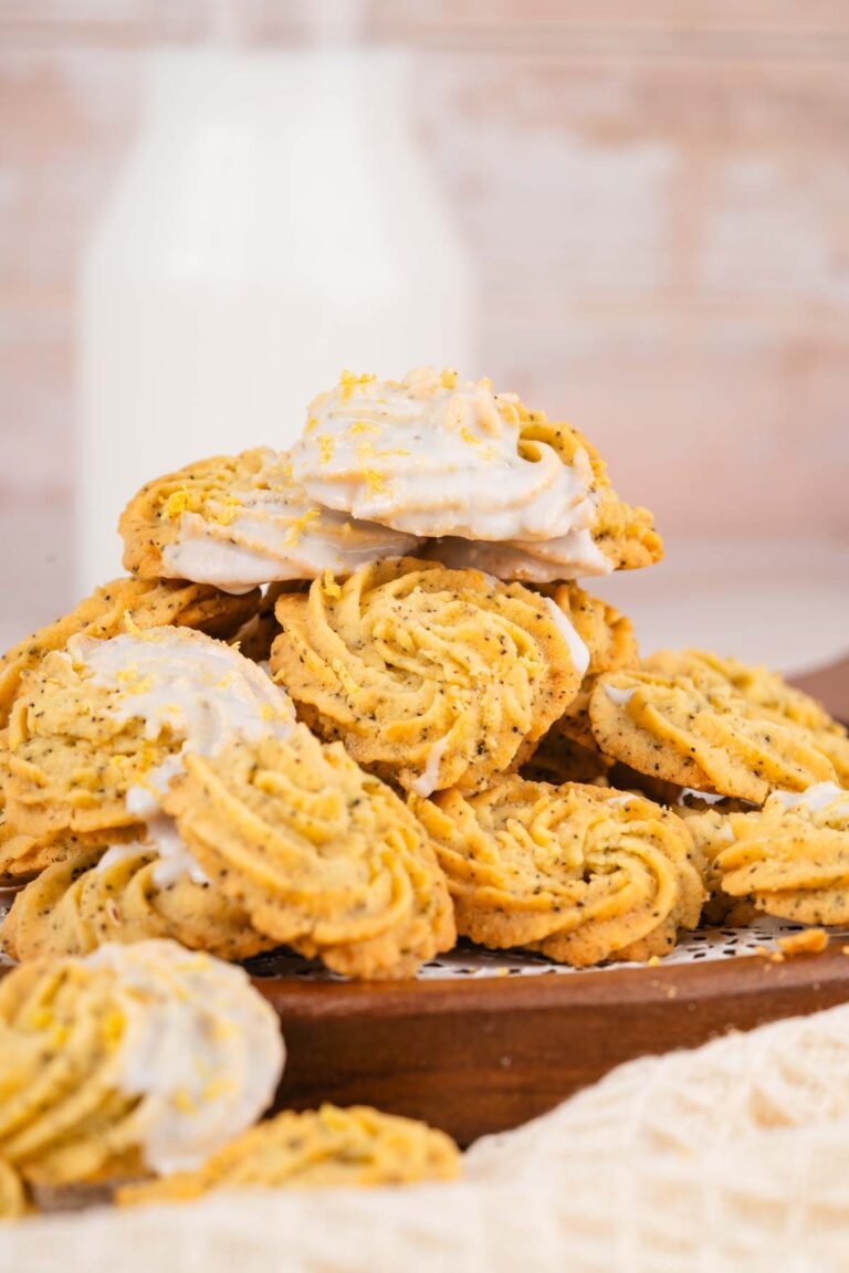 A pile of swirled Earl Grey Lemon Cookies with poppy seeds, white icing, and lemon zest sits on a wooden tray, with a bottle of milk in the background.