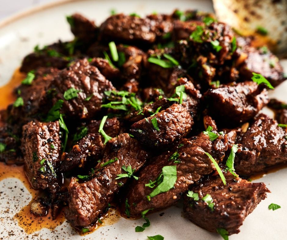 Cooked beef tips, inspired by Garlic Butter Steak Bites, garnished with chopped fresh herbs on a plate, with visible juices and seasoning.