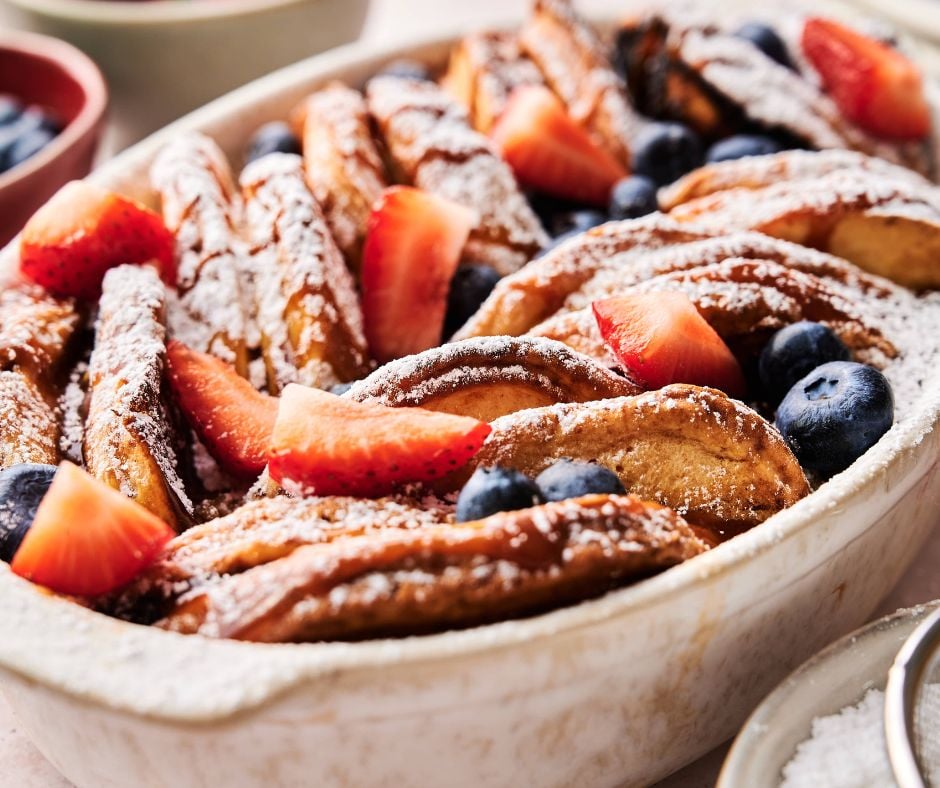 A baked pancake casserole of sliced French toast arranged in a dish, topped with powdered sugar, fresh strawberries, and blueberries.
