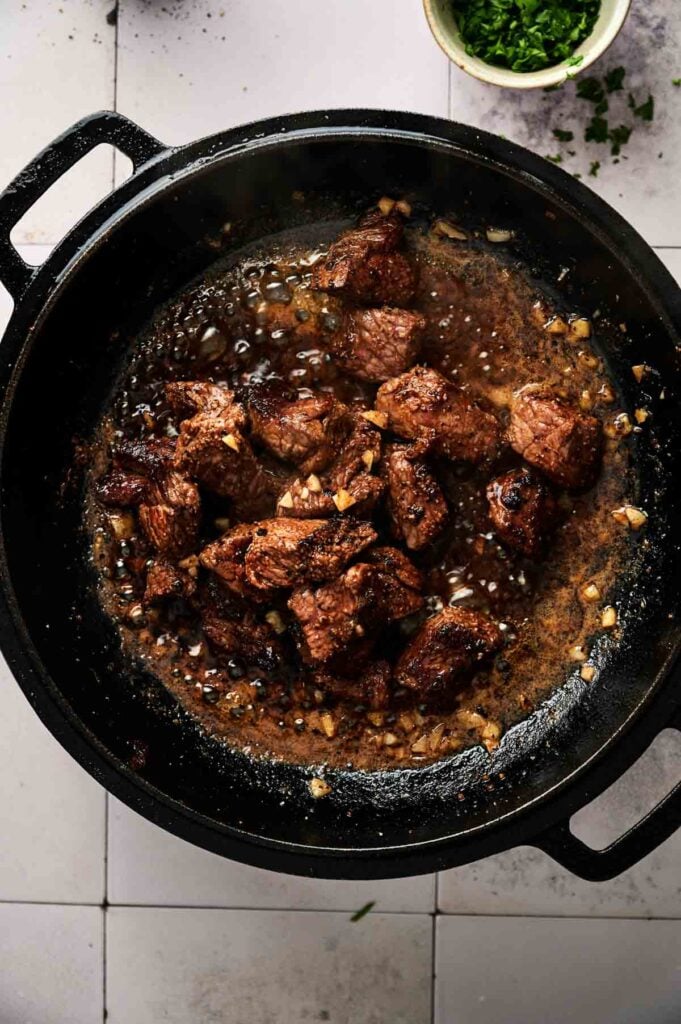 Chunks of beef are searing in a cast iron pan with diced garlic and sauce, creating irresistible Garlic Butter Steak Bites, with chopped herbs visible in a bowl nearby.