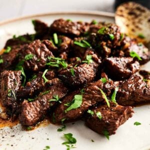 Garlic Butter Steak Bites served on a white plate, garnished with chopped parsley, with a spoon and pepper grinder in the background.