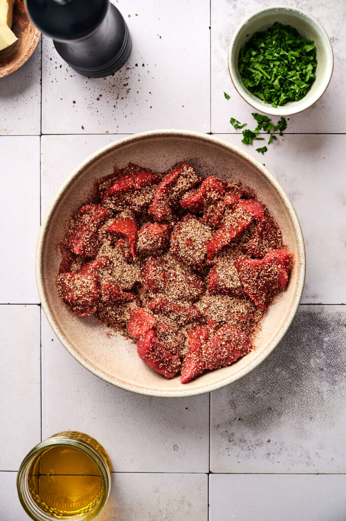 A bowl of raw beef pieces coated with spices, ready to become Garlic Butter Steak Bites, is surrounded by chopped herbs in a small bowl, a bottle of oil, and a pepper grinder on a white tiled surface.