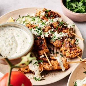 A plate of Greek chicken kebobs topped with sauce and chopped herbs, served with a bowl of creamy dip and a side of fresh parsley; a tomato is in the foreground.