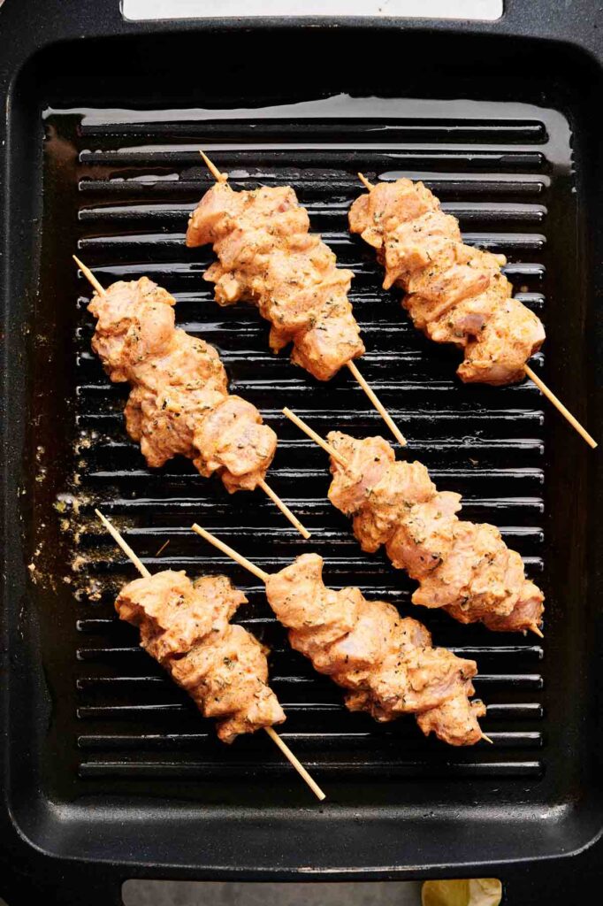 Five raw, marinated Greek chicken kebobs are arranged on a black grill pan, ready to be cooked.