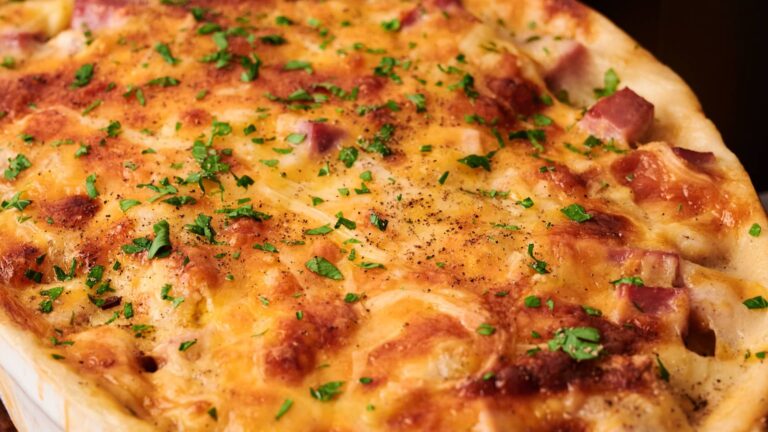 A baked casserole topped with melted cheese, diced ham, and chopped parsley, showing a golden-brown crust.