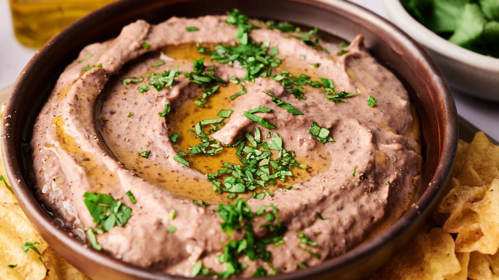A bowl of bean dip garnished with chopped herbs and olive oil, surrounded by tortilla chips.
