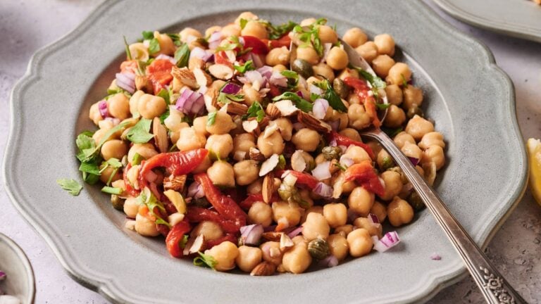 A plate of chickpea salad with chopped red onion, roasted red peppers, capers, almonds, and fresh herbs, served with a spoon.