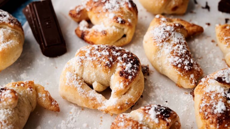 Several small, crescent-shaped pastries dusted with powdered sugar are arranged on parchment paper next to pieces of chocolate.