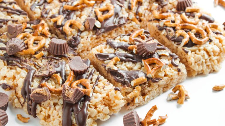 Rice crispy treats topped with mini peanut butter cups, pretzel pieces, and a drizzle of chocolate, cut into squares and arranged on a white surface.