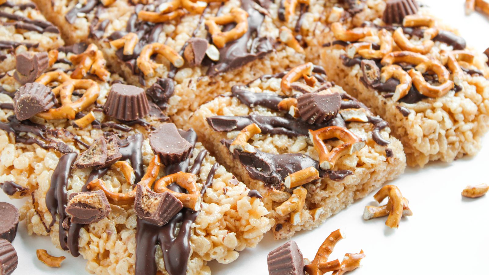 Rice crispy treat bars topped with drizzled chocolate, mini peanut butter cups, and crushed pretzels, cut into squares and arranged on a white surface.