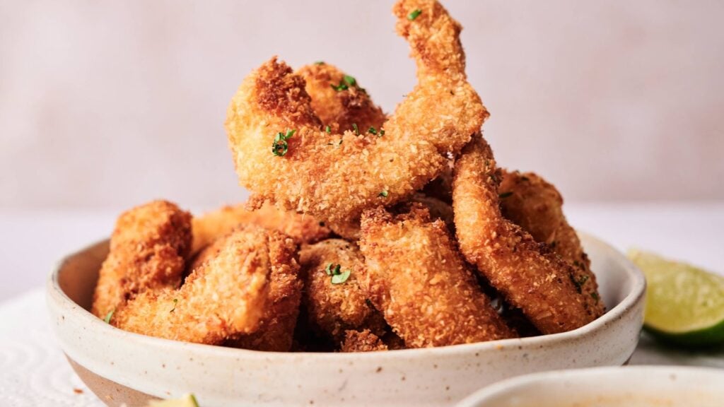 A bowl filled with golden-brown breaded shrimp garnished with chopped herbs, with a wedge of lime visible on the side.