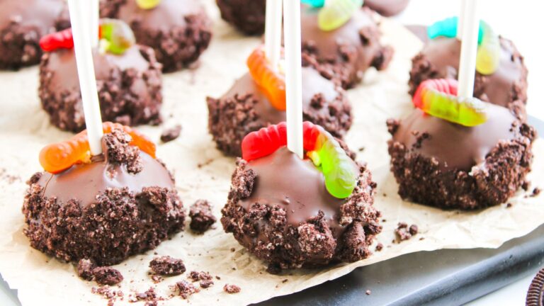 Chocolate-covered cake pops topped with gummy worms and crushed cookies, arranged on parchment paper.