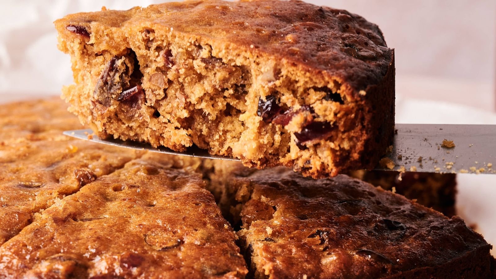 A close-up of a fruitcake with a slice being lifted out, showing its dense texture and pieces of dried fruit inside.