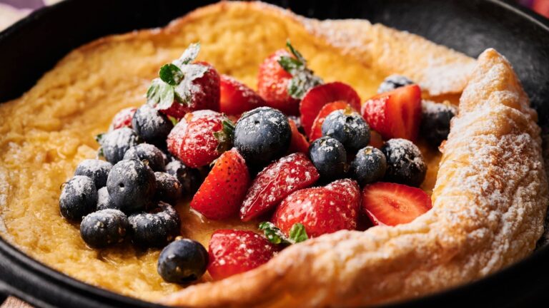 A Dutch baby pancake topped with fresh strawberries, blueberries, and a dusting of powdered sugar, served in a skillet.