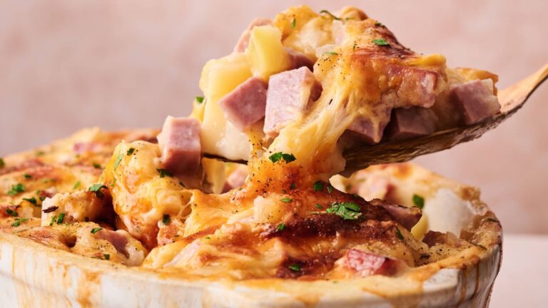 A close-up of a cheesy casserole with chunks of ham and potatoes, being served with a wooden spoon.