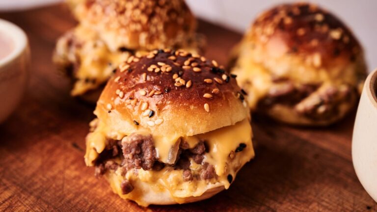 Three sesame seed slider buns filled with ground meat and melted cheese are arranged on a wooden surface.