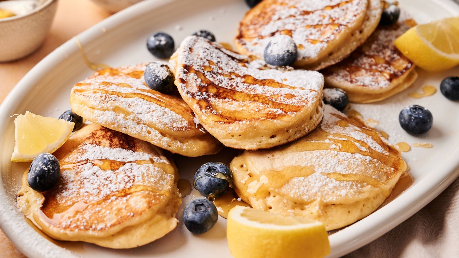 A plate of pancakes topped with powdered sugar, fresh blueberries, lemon wedges, and drizzled with syrup.