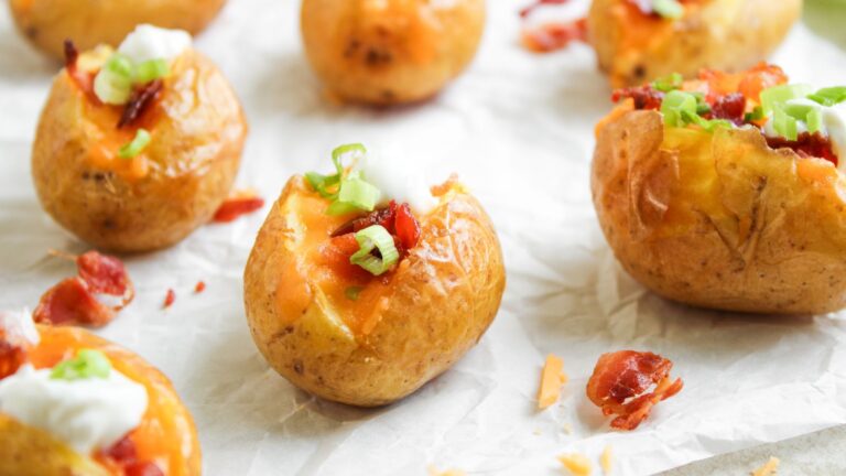 Small baked potatoes topped with melted cheese, bacon bits, sour cream, and chopped green onions on parchment paper.